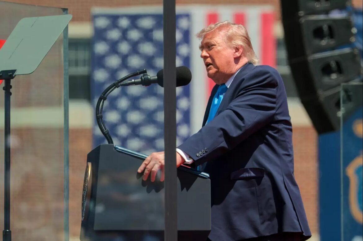 Man in a dark suit and blue tie speaks at an outdoor podium, microphone in front, with a blurred American flag backdrop.