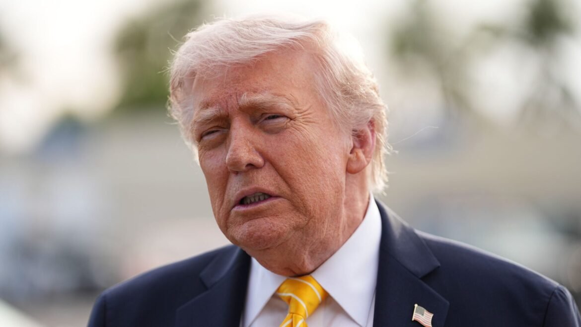 Close-up of an older man in a navy suit and yellow striped tie outdoors, with a blurred background.