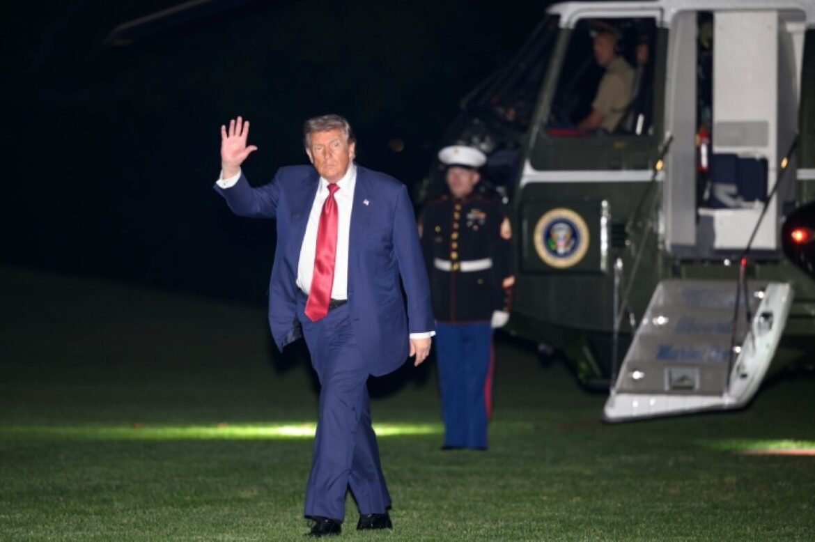 A man in a navy suit and red tie waving as he walks on a grassy tarmac near a large helicopter with guards nearby.