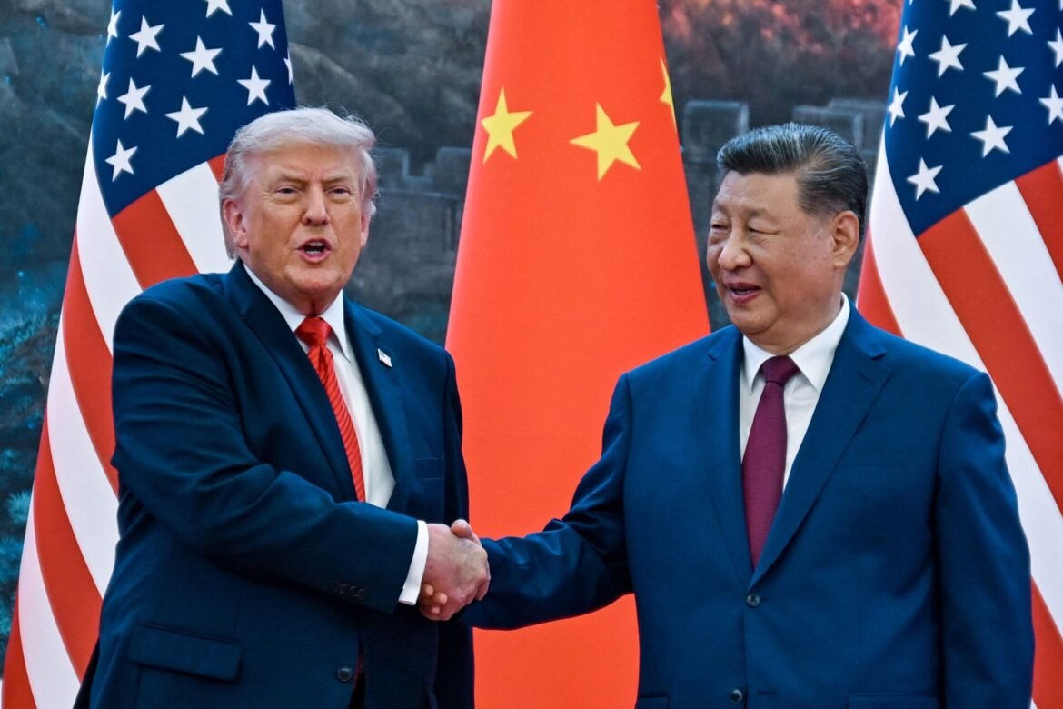 Two men in dark suits shake hands in front of U.S. and Chinese flags at a formal event.