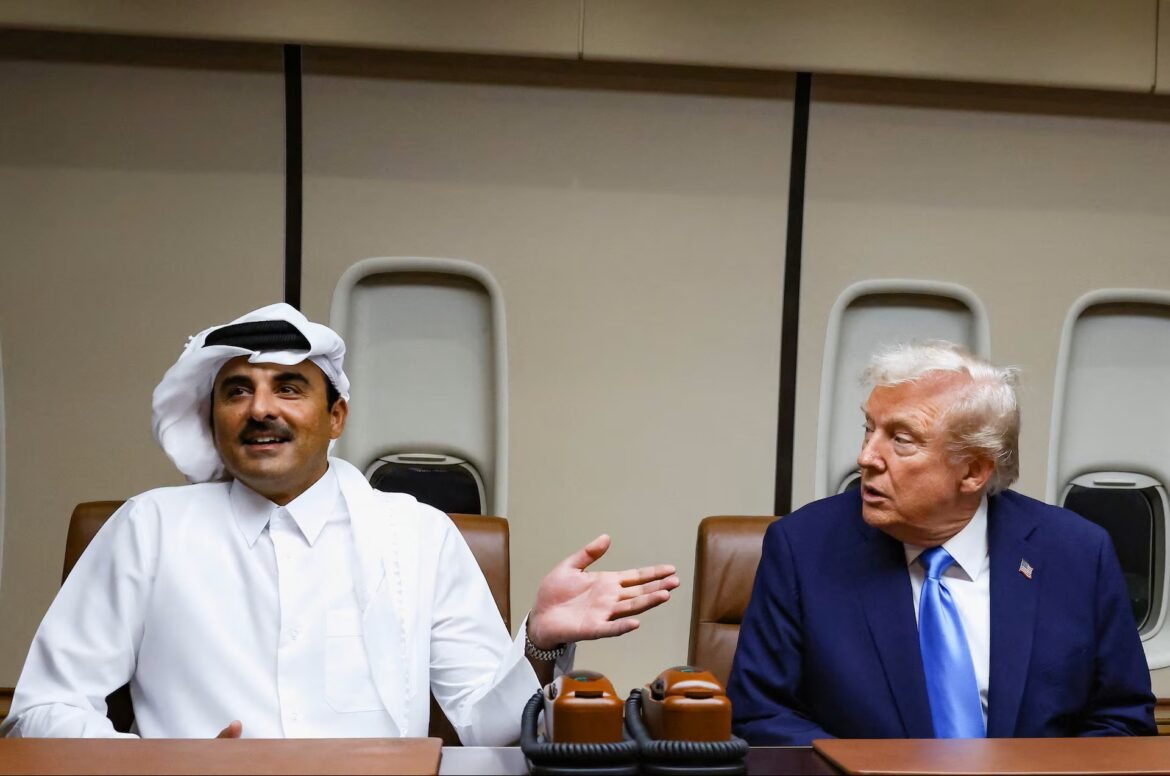 Two men sit in an airplane cabin at a conference table, one in white traditional dress gesturing as the other in a navy suit listens.