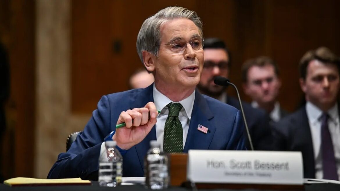 An older man in a navy suit and green tie testifies at a formal hearing, speaking into a microphone with a nameplate in front.