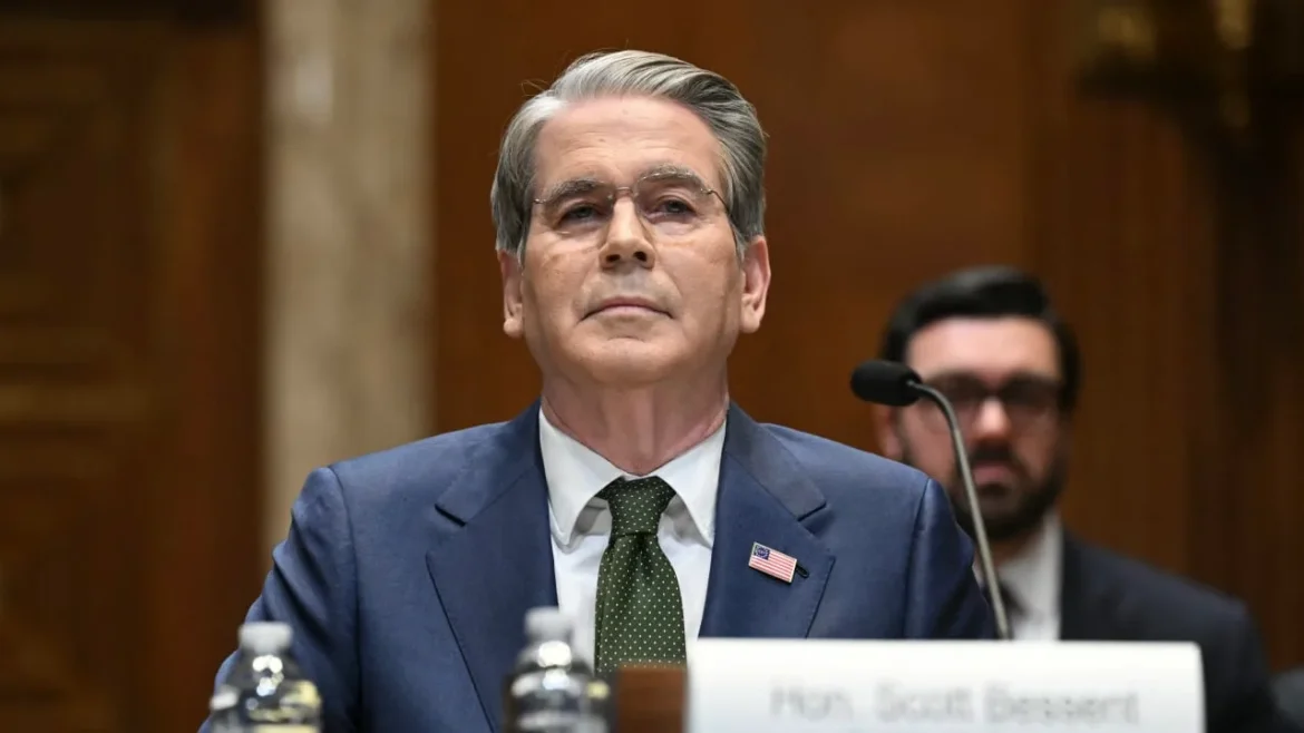 Middle-aged man in a blue suit and green tie sits at a panel desk with a microphone, wearing a small U.S. flag pin in a formal hearing room.