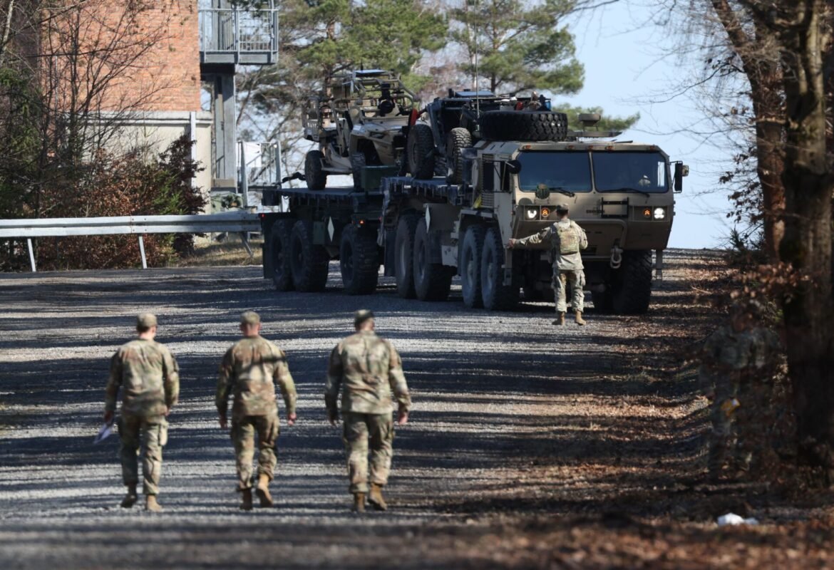 Soldiers in camouflage observe a convoy of armored vehicles and jeeps loaded on a large transport truck on a tree-lined road.