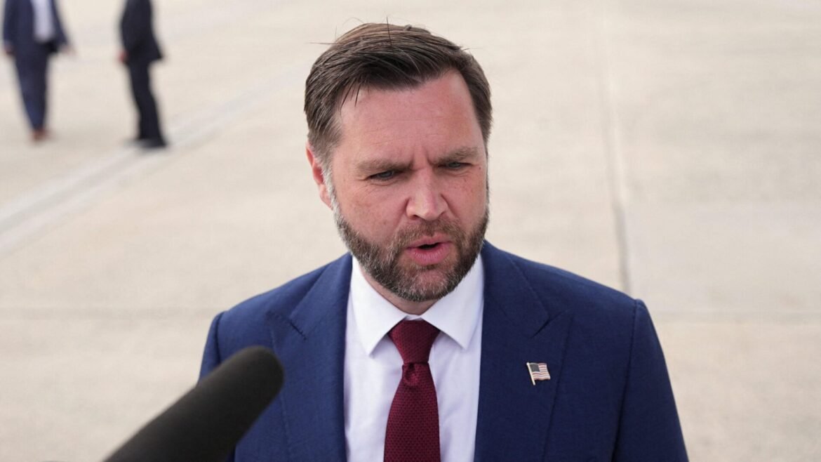 Man in a navy suit and red tie speaks to reporters outdoors, a microphone visible in the foreground and an American flag pin on his lapel.