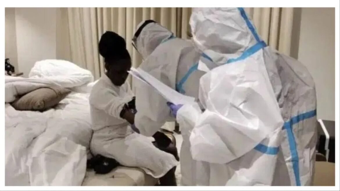Healthcare worker in full protective gear assists a woman seated on a bed in a clinical room during care or testing.