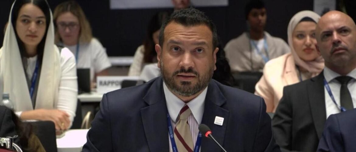 Man in a dark suit and striped tie speaks at a conference, microphone in front, with attendees in the background.