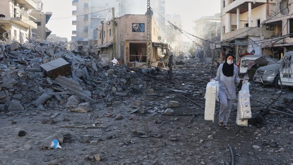 Woman in hijab wearing a mask carrying large bags of supplies through a rubble-strewn street after buildings were destroyed.