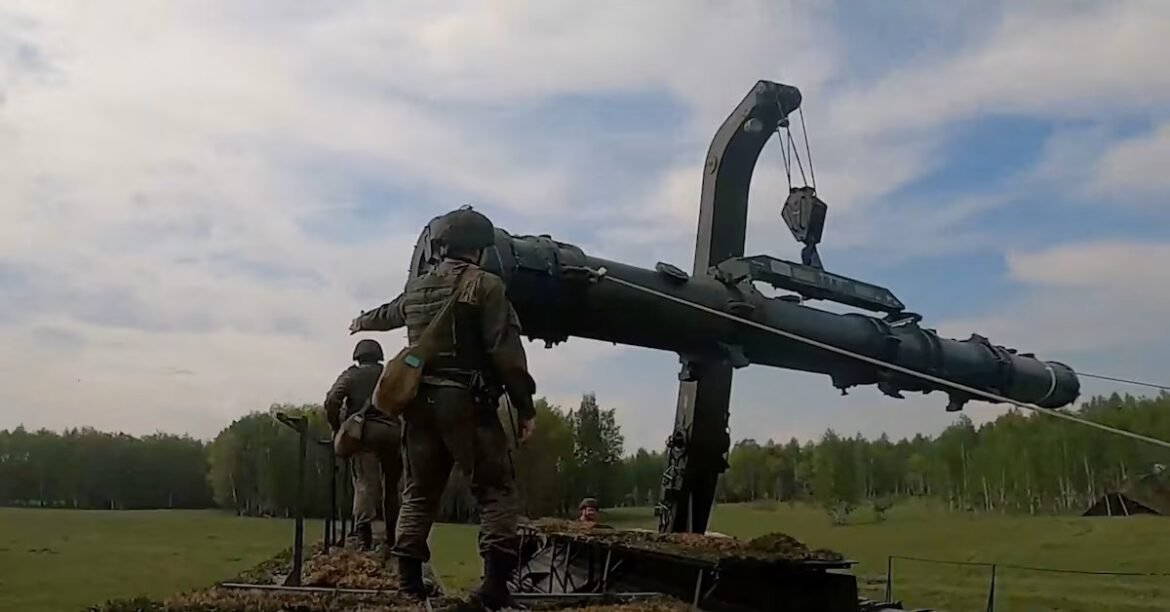 Soldiers in uniform stand beside a large field artillery gun with a crane lifting a shell in a grassy, open area.