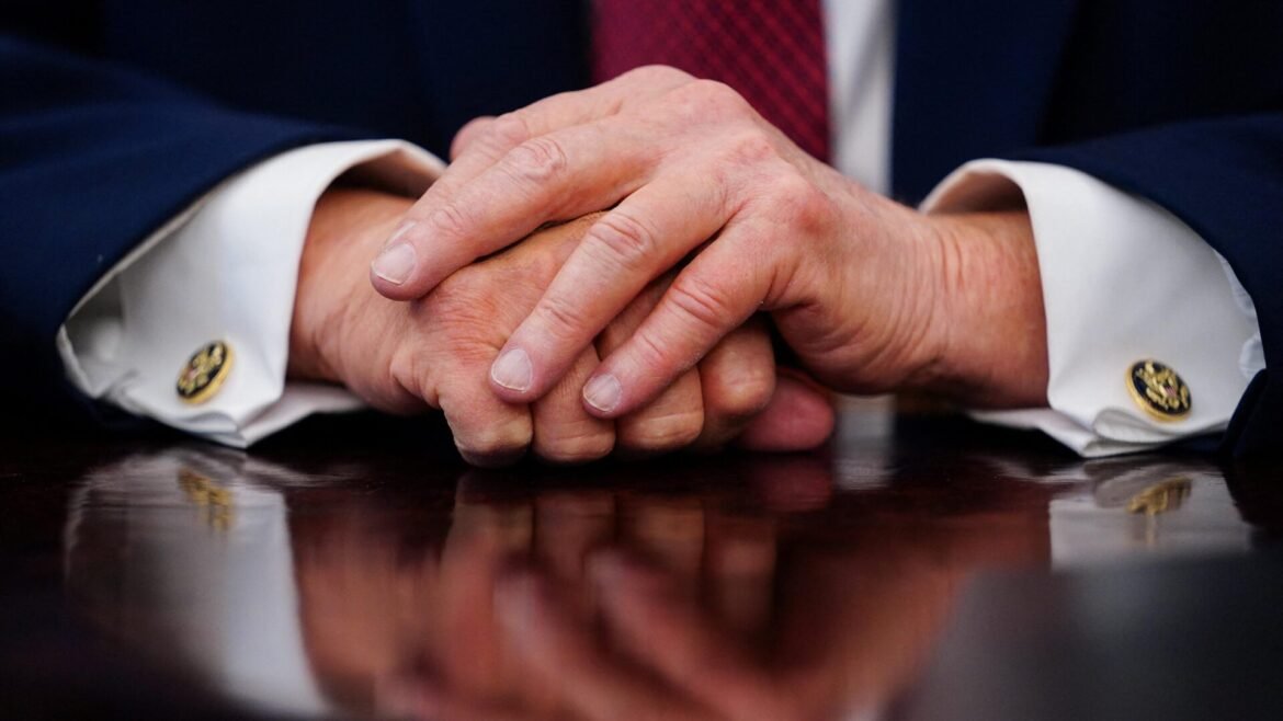 Two hands clasped on a dark wooden table, wearing a navy suit and white cuffs with gold cufflinks.