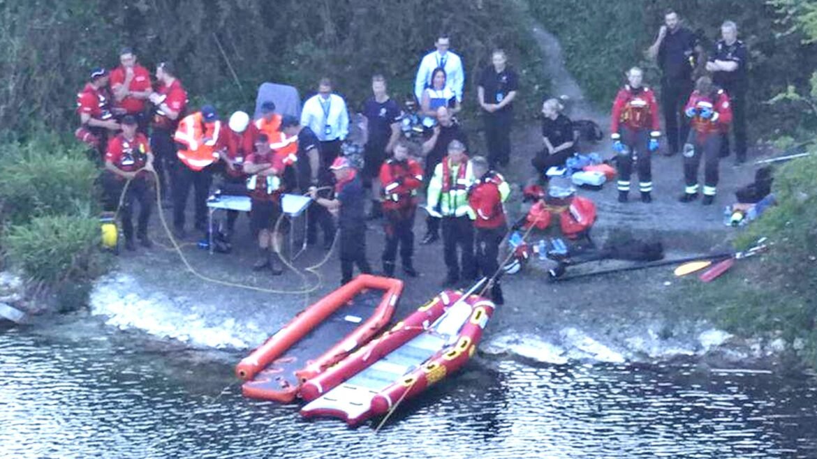 Emergency responders in red uniforms and life jackets gather on a riverbank with inflatable rescue boats nearby.