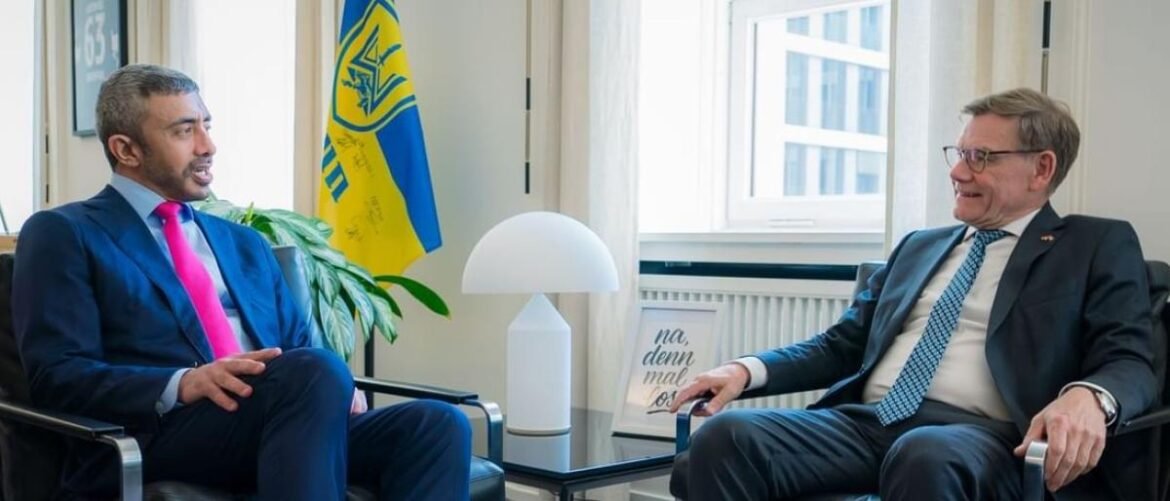 Two men in suits sit in chairs facing each other and talk in a bright office, with a yellow-and-blue flag in the background and a white lamp nearby.