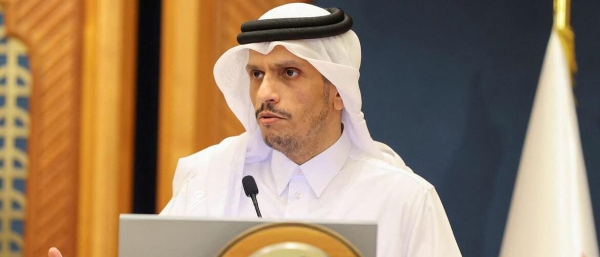 Man wearing a white thobe and headscarf speaks at a podium with a microphone in a formal setting.