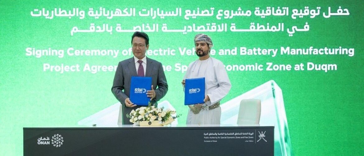 Two men in formal attire and traditional Omani dress hold blue folders during a signing ceremony at a green backdrop announcing an electric vehicle and battery project in Duqm.