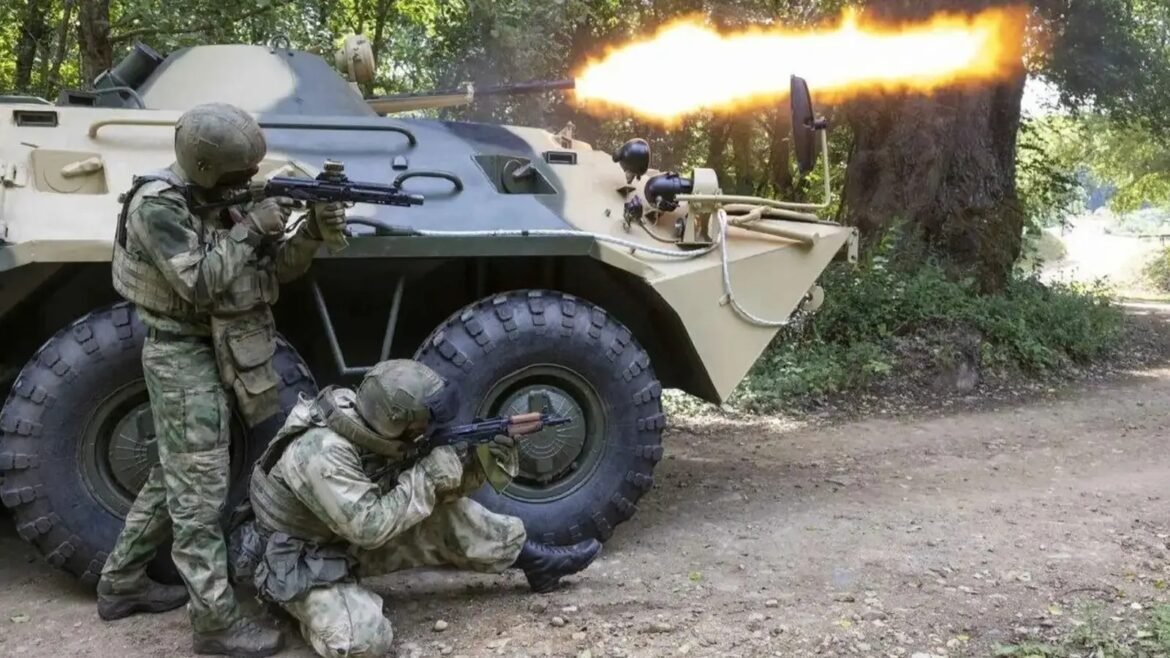Two soldiers in camouflage fire a mounted machine gun beside an armored vehicle in a wooded area, with a muzzle flash visible.