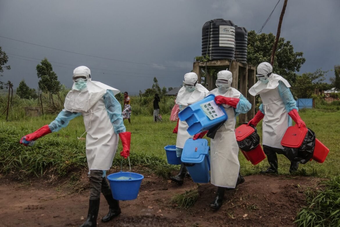 Health workers in white hazmat suits and red gloves carrying blue and red sample containers in a muddy field.
