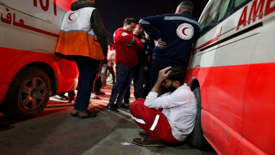 Man seated on the ground with head in hands beside a red-and-white ambulance, while rescuers and volunteers stand nearby in uniforms.