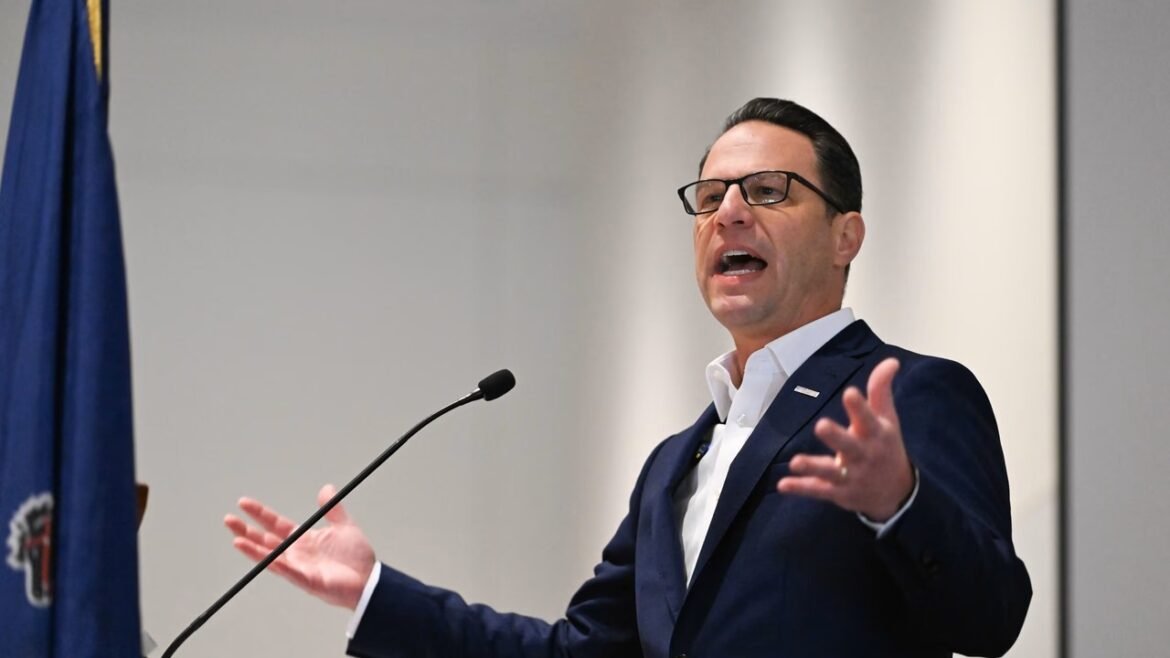 Man in a navy suit and glasses speaks at a podium with a microphone during a formal event in front of a flag.