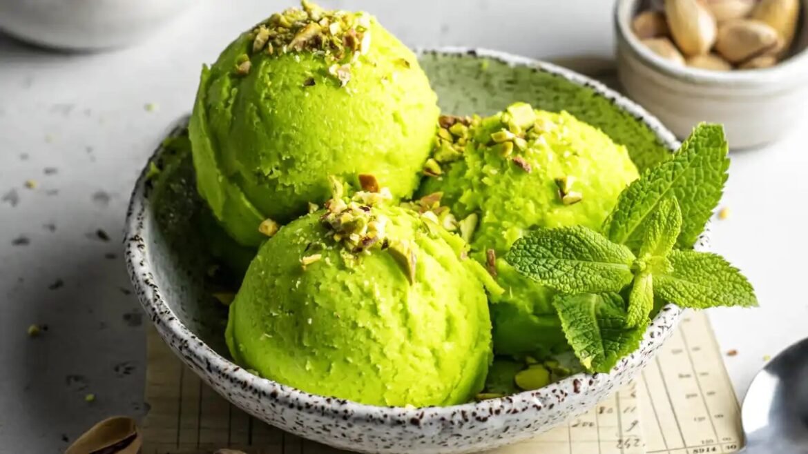 Three scoops of bright green pistachio ice cream topped with chopped pistachios and a sprig of mint in a speckled bowl.