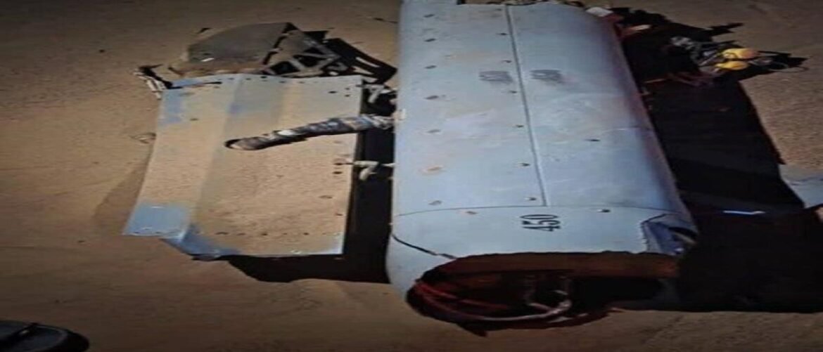 Damaged grey aircraft fuselage panel and cylindrical section on a dusty ground, with a bent pipe and scattered debris.
