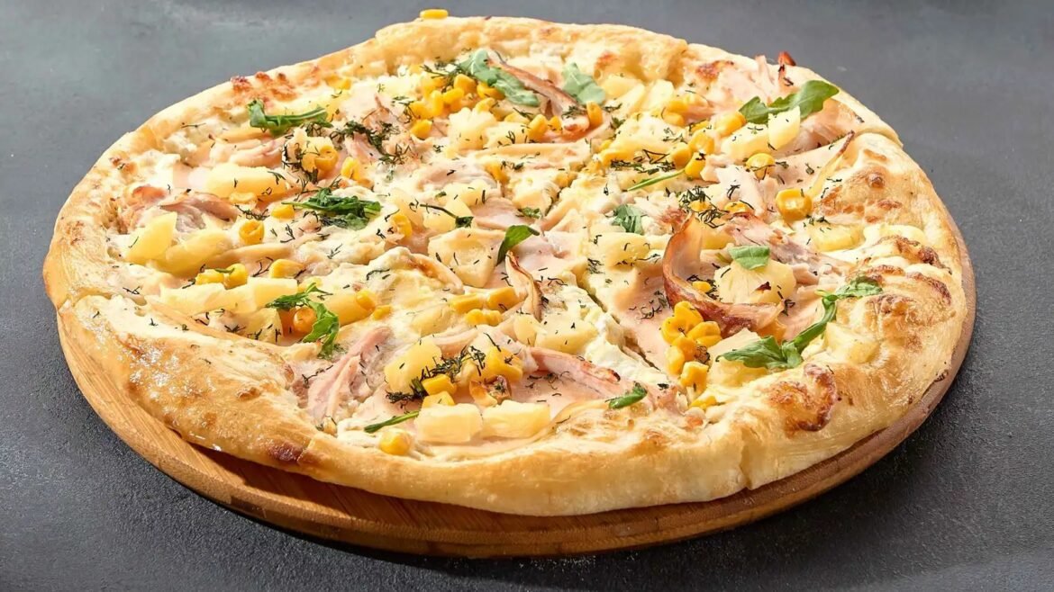 Pizza topped with pineapple, corn, ham, and herbs on a golden crust, served on a wooden board