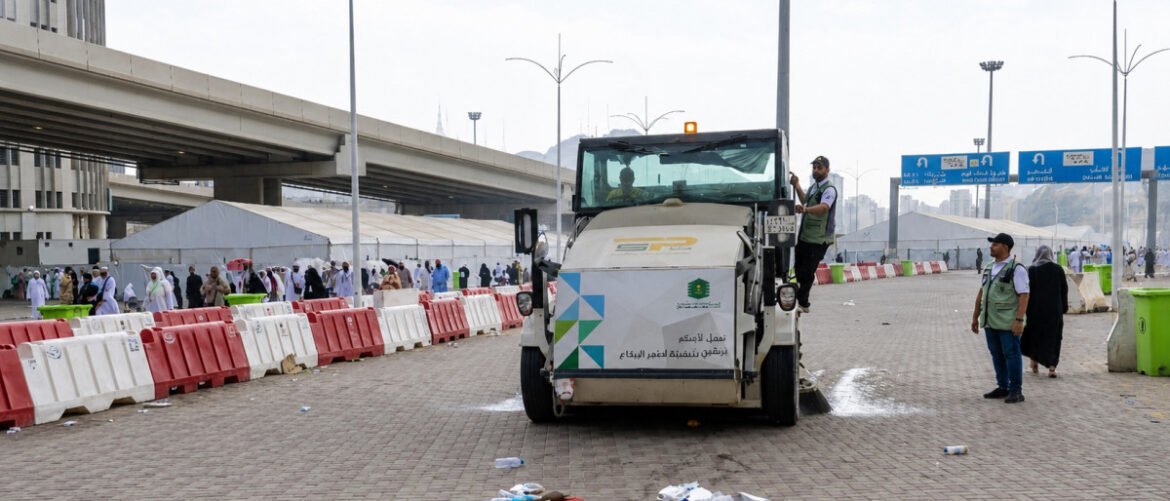 A street-sweeping truck operates in a plaza bordered by red and white barriers while workers in vests guide it and people walk nearby under an overpass ground tents visible in the background.