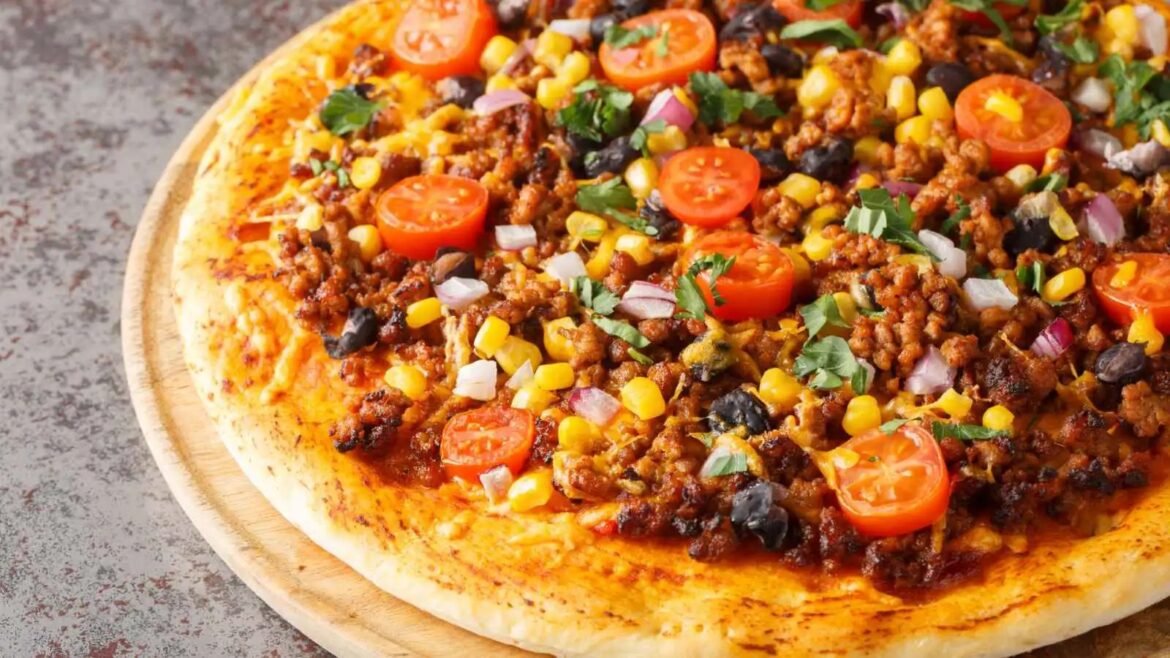 Pizza topped with cherry tomatoes, corn, onions, olives, cilantro, and seasoned ground meat on a gold-brown crust on a wooden board.