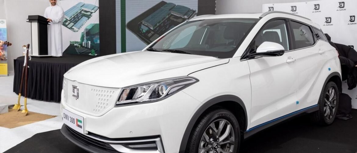 White crossover SUV on a display stage at an auto show, with a podium and presenter in the background.