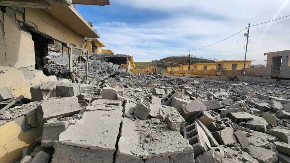 Rubble-strewn street with collapsed concrete blocks and damaged walls; yellow houses intact in distance under a blue sky.