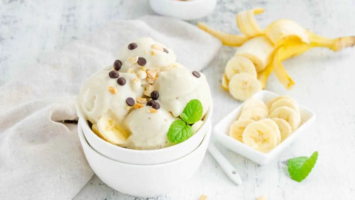 Banana ice cream scoops in a white bowl topped with chocolate chips and nuts, with fresh mint and banana slices on the side.