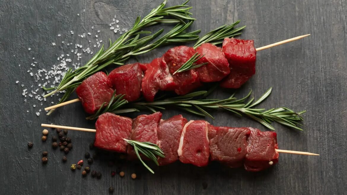 Raw beef skewers with fresh rosemary on a dark surface, sprinkled with coarse salt and peppercorns.