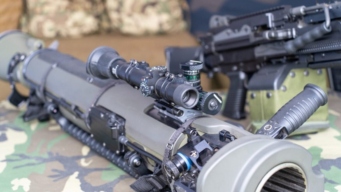 Close-up of a sniper rifle with a scope and bipod resting on camouflage-patterned ground in a field setting.