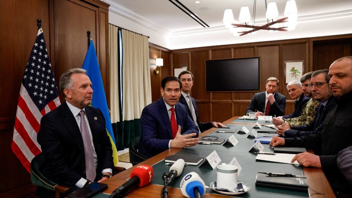 Diplomatic meeting around a long conference table with US and Ukrainian flags in the background on a wood-paneled wall.