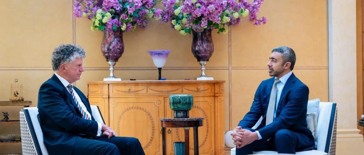 Two men in suits sit facing each other in a formal room, discussing, with a carved wooden cabinet and large purple flower arrangements behind them.