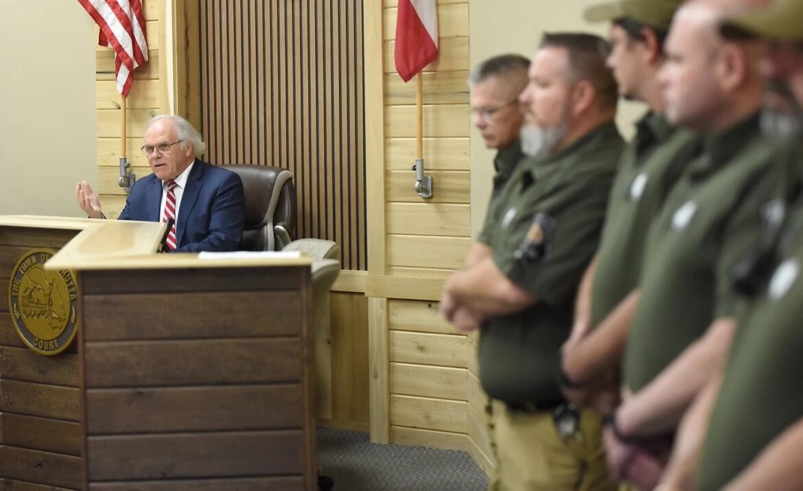 Judge at a courtroom bench speaks, gesturing with one hand while uniformed officers stand in a line to the right, flags in the background.