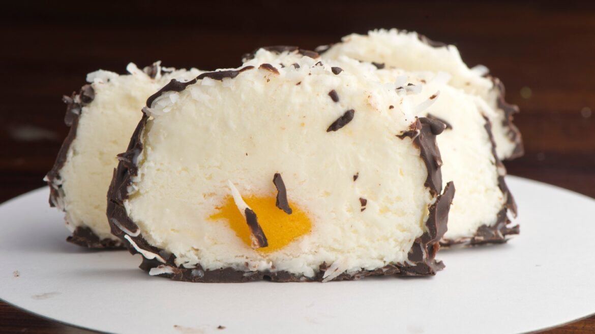 Slice of white chocolate-coated ice cream bomb with orange center on a white plate, dark chocolate shavings sprinkled over it