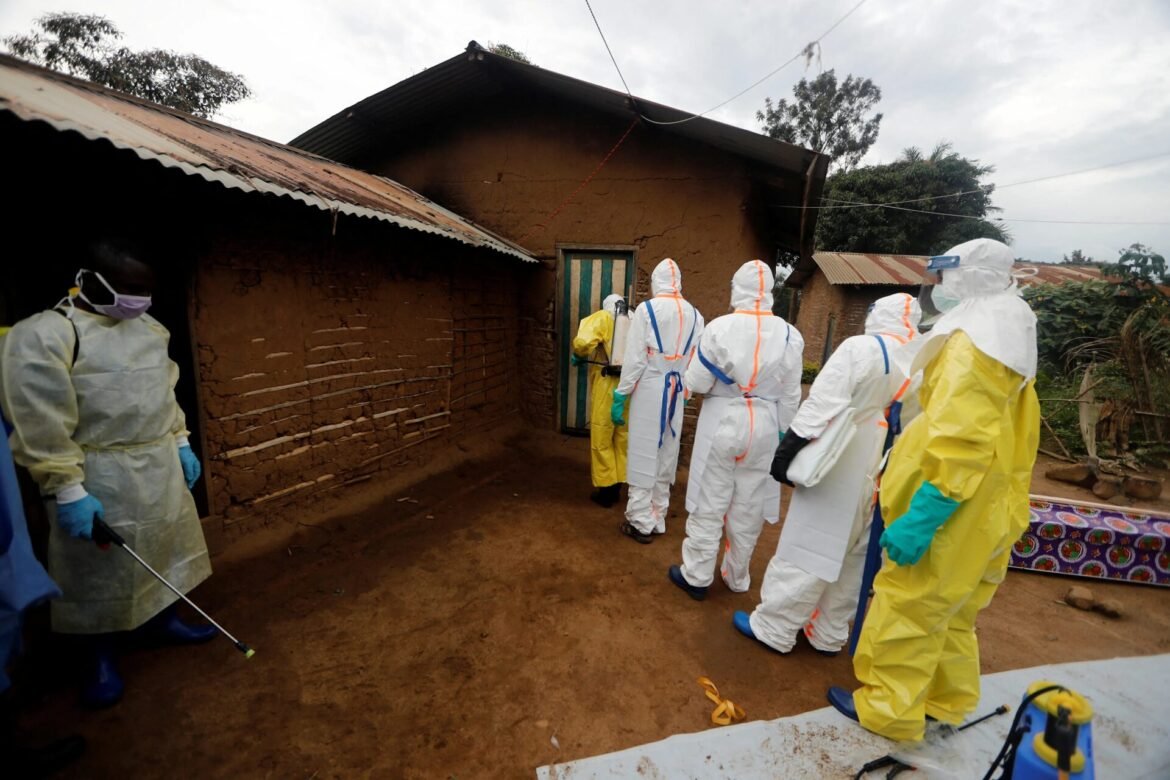 Group of people in full PPE suits and masks outside a mud brick rural house, carrying spraying equipment.