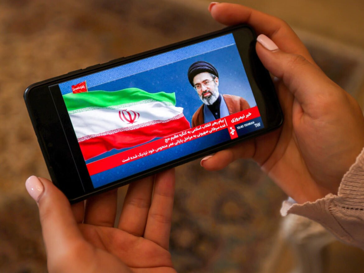 Handheld smartphone showing a Persian news broadcast with the Iranian flag and a cleric against a blue background. A red ticker runs along the bottom in Persian text.