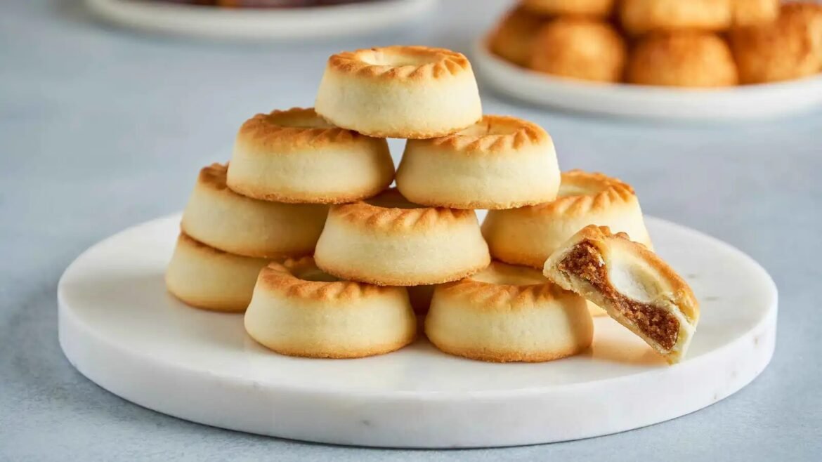Stack of small round shortbread cookies arranged in a pyramid on a white pedestal, with one cookie broken open to reveal filling.
