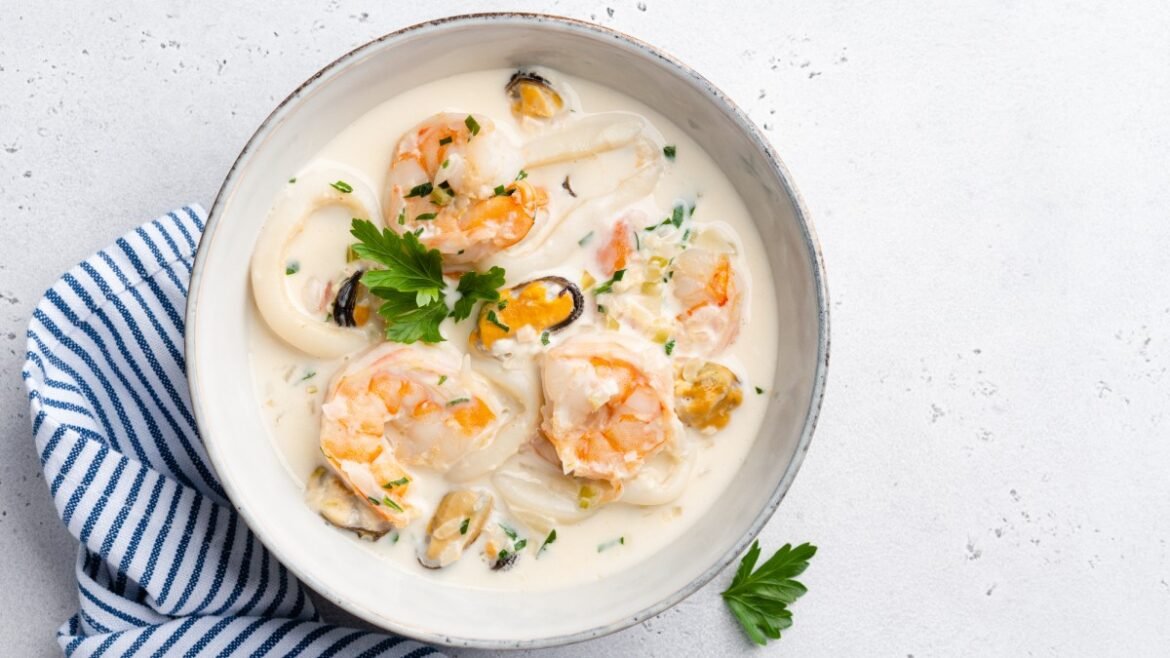 Creamy seafood chowder with shrimp, mussels, and parsley in a white bowl on a pale surface with a blue striped napkin nearby.