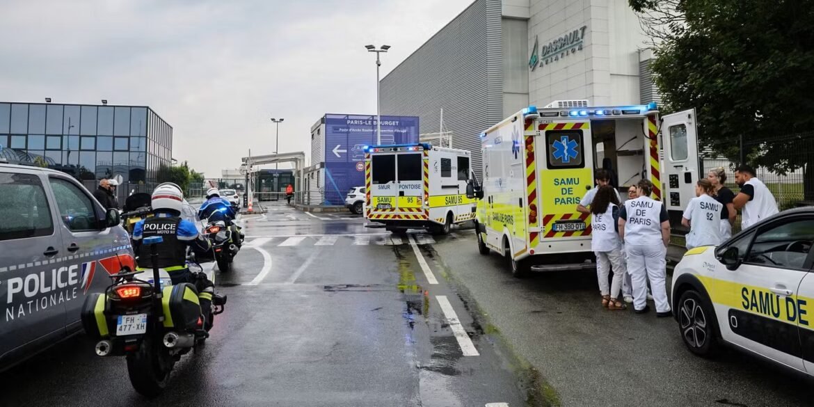 Emergency responders and ambulances outside a Dassault Aviation building; police car, motorcycles, and EMS crews with SAMU de Paris vests on a wet street edge.