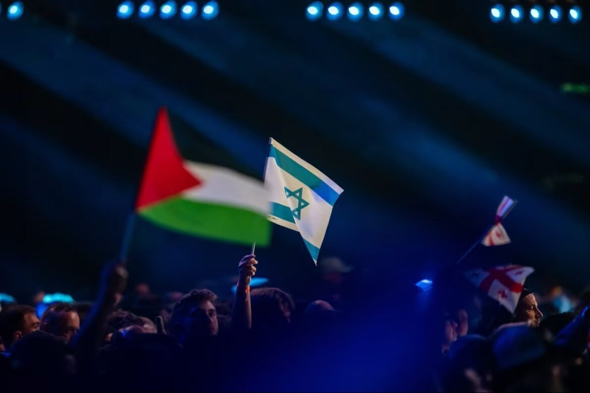 Crowd at a nighttime rally waving Israeli and Palestinian flags under blue stage lights.