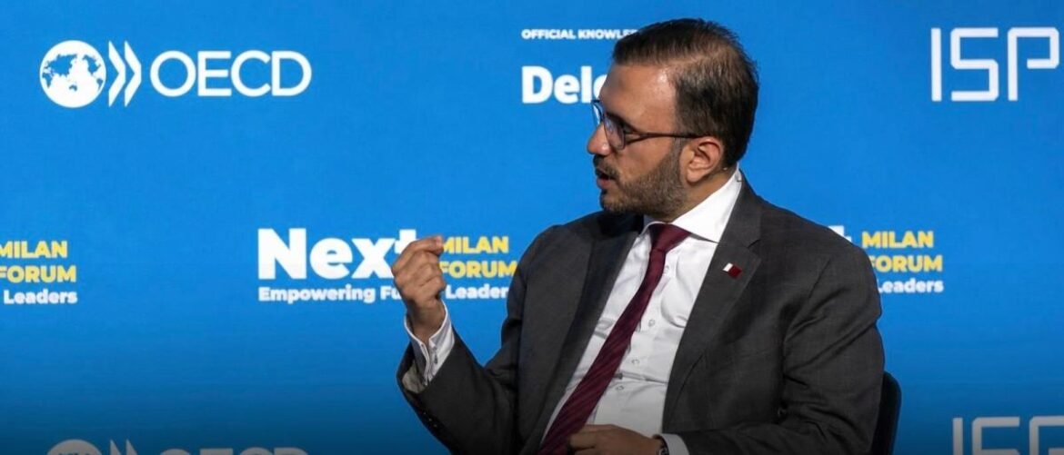 Man in a gray suit and red tie speaks on stage, gesturing with his right hand, against a blue backdrop with OECD and Milan Forum logos on display.