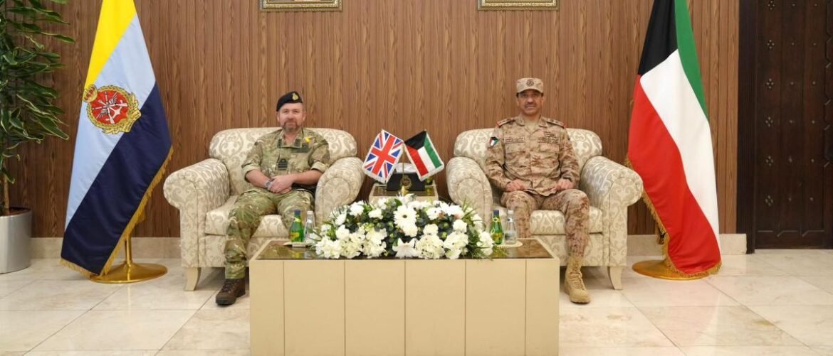 Two uniformed officers sit in beige armchairs for a formal military meeting, with flags and a flower centerpiece between them.