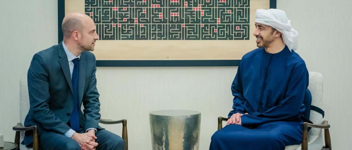 Two professionals in a formal meeting: a man in a blue suit and a man in traditional Middle Eastern dress sit facing each other in a modern office setting.