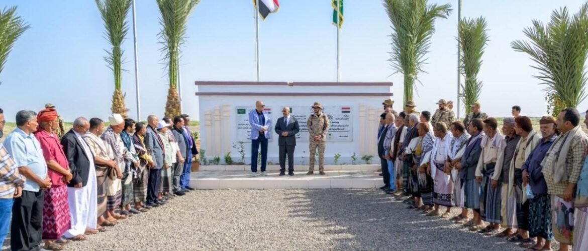 Group of men and women standing in two lines facing a white memorial wall with a panel, during an outdoor ceremony with flags in the background.