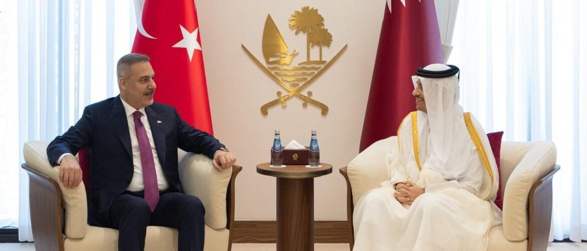 Two men in formal attire sit in cream armchairs facing each other in a diplomatic meeting, with Turkish and Qatari flags nearby and a gold emblem on the wall behind.