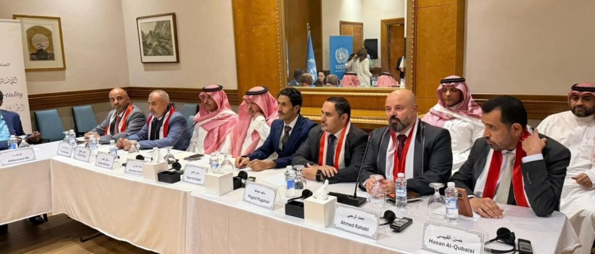 Panel of men in formal attire with red scarves seated at a long white-clothed conference table in a meeting room; nameplates, microphones, water bottles, and a UN banner visible in the background.