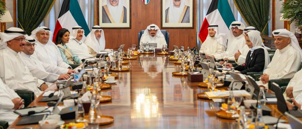 Diplomatic officials in traditional dress seated around a long conference table with laptops and microphones, Kuwait flags and portraits in the background.