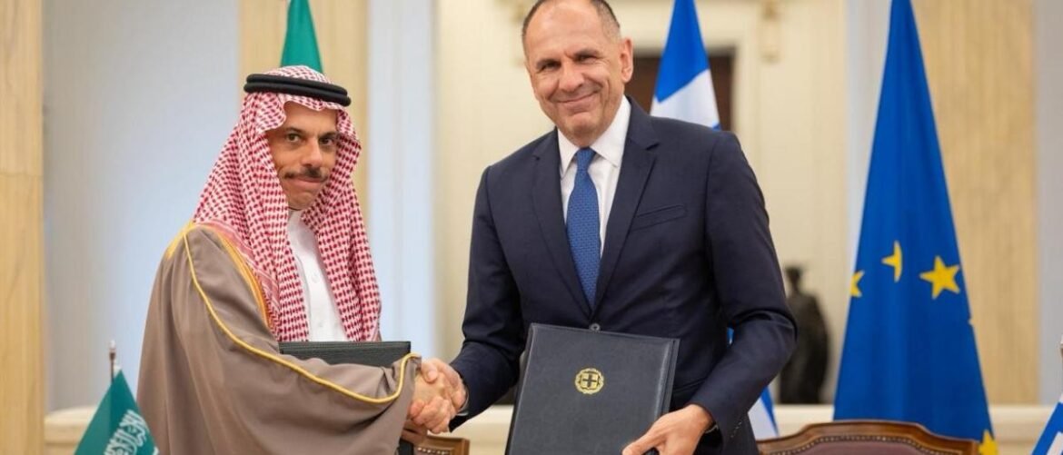 Two men in formal attire shake hands while holding folders at a signing ceremony, with flags in the background.